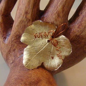 May include: A gold-colored leaf-shaped ornament with copper wire detailing. The leaf has visible veins and a textured surface. The copper wire is coiled and adds a decorative element to the leaf. The ornament is likely a decorative item.