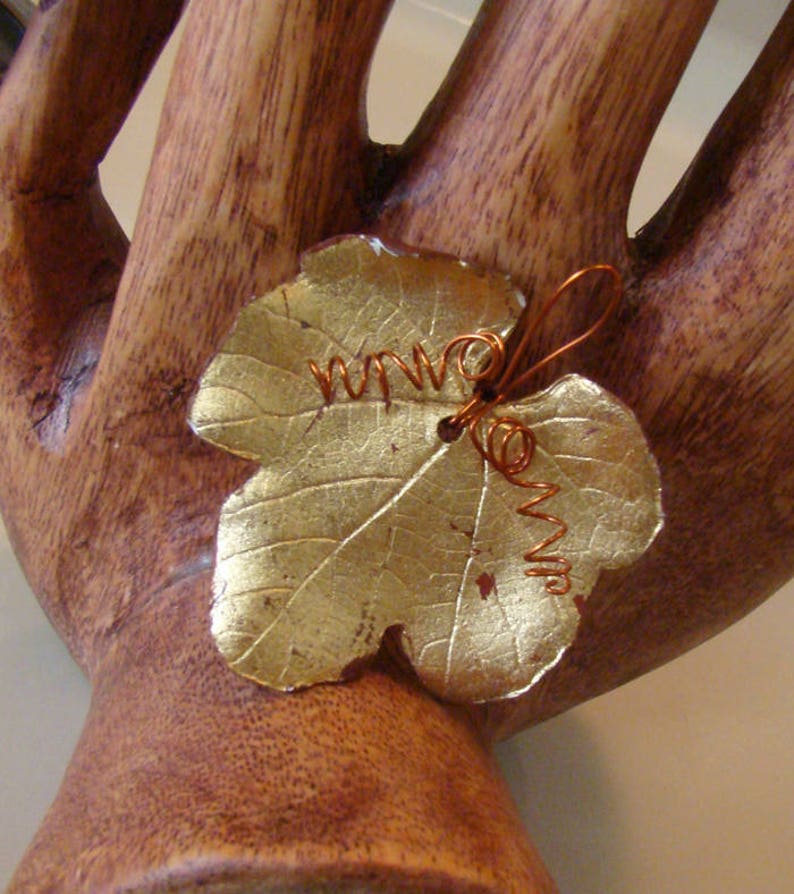 May include: A gold-colored leaf-shaped ornament with copper wire detailing. The leaf has visible veins and a textured surface. The copper wire is coiled and adds a decorative element to the leaf. The ornament is likely a decorative item.