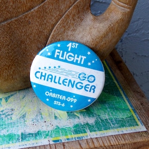 May include: A blue and white button with the text "1st Flight GO CHALLENGER ORBITER-099 STS-6" and stars around the edge.