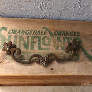May include: Wooden box with the words "Orangedale Oranges Sunflower" in green lettering. A decorative bronze-colored handle with floral accents is attached to the front of the box.