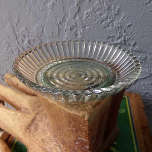 May include: A clear glass dish with a fluted edge sits on top of a wooden hand sculpture. The dish has a circular design in the center.