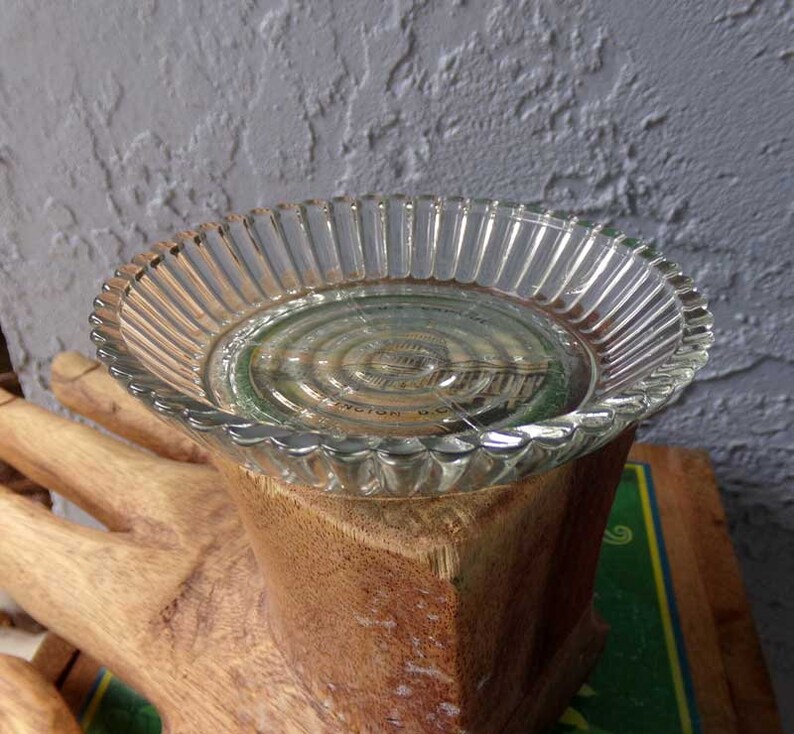 May include: A clear glass dish with a fluted edge sits on top of a wooden hand sculpture. The dish has a circular design in the center.