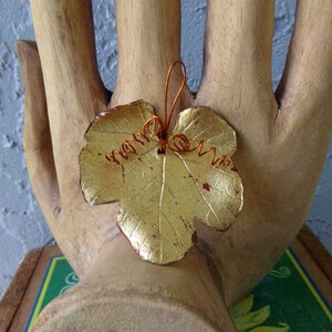 May include: A gold-colored leaf-shaped pendant with copper wire detailing. The leaf has a textured surface and is attached to a copper wire stem. The pendant is displayed on a wooden hand sculpture.