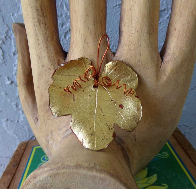 May include: A gold-colored leaf-shaped pendant with copper wire detailing. The leaf has a textured surface and is attached to a copper wire stem. The pendant is displayed on a wooden hand sculpture.