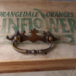 May include: A vintage bronze drawer pull with ornate detailing is mounted on a wooden surface. The wood has faded green lettering that reads "ORANGEDALE ORANGES SUNFLOWER". The drawer pull has a curved handle and decorative accents.