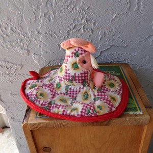 May include: A red, white, and green fabric chicken with a sunflower and gingham pattern. The chicken is sitting on a wooden box.
