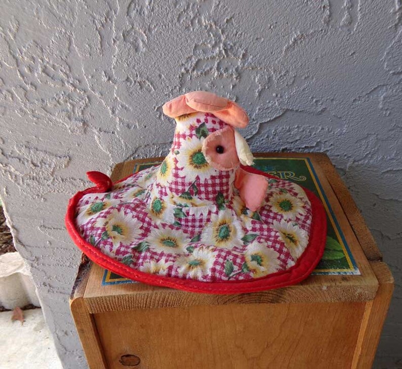 May include: A red, white, and green fabric chicken with a sunflower and gingham pattern. The chicken is sitting on a wooden box.