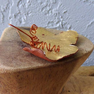 May include: A handcrafted leaf-shaped brooch with a gold-colored surface and a copper wire accent. The leaf is painted with gold and red tones. The brooch is resting on a wooden surface.