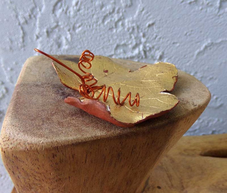 May include: A handcrafted leaf-shaped brooch with a gold-colored surface and a copper wire accent. The leaf is painted with gold and red tones. The brooch is resting on a wooden surface.