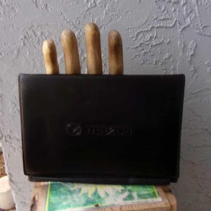 May include: A black leather knife holder with four wooden-handled knives. The holder has the Mazda logo embossed on the front. The knives are visible above the holder's top edge. The holder is sitting on a wooden surface.