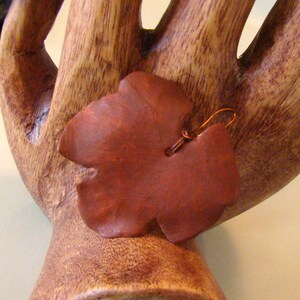 May include: A reddish-brown leaf-shaped earring with a copper-colored wire hook. The earring is made of a smooth, possibly wooden material. The earring is displayed on a wooden hand-shaped sculpture.