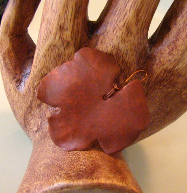 May include: A reddish-brown leaf-shaped earring with a copper-colored wire hook. The earring is made of a smooth, possibly wooden material. The earring is displayed on a wooden hand-shaped sculpture.