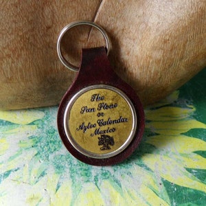 May include: A brown leather keychain with a gold-colored metal circle featuring the text "The Sun Stone or Aztec Calendar Mexico".