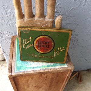May include: Vintage Lucky Strike cigarettes tin from the fifties. The rectangular tin is green with a red and yellow circular logo. The words "Lucky Strike" and "Cigarettes" are visible. The tin is placed on a wooden surface.