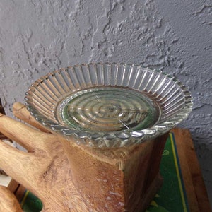 May include: A clear glass dish with a fluted edge sits on top of a wooden hand sculpture. The dish has a circular design with a faint image of a building in the center.