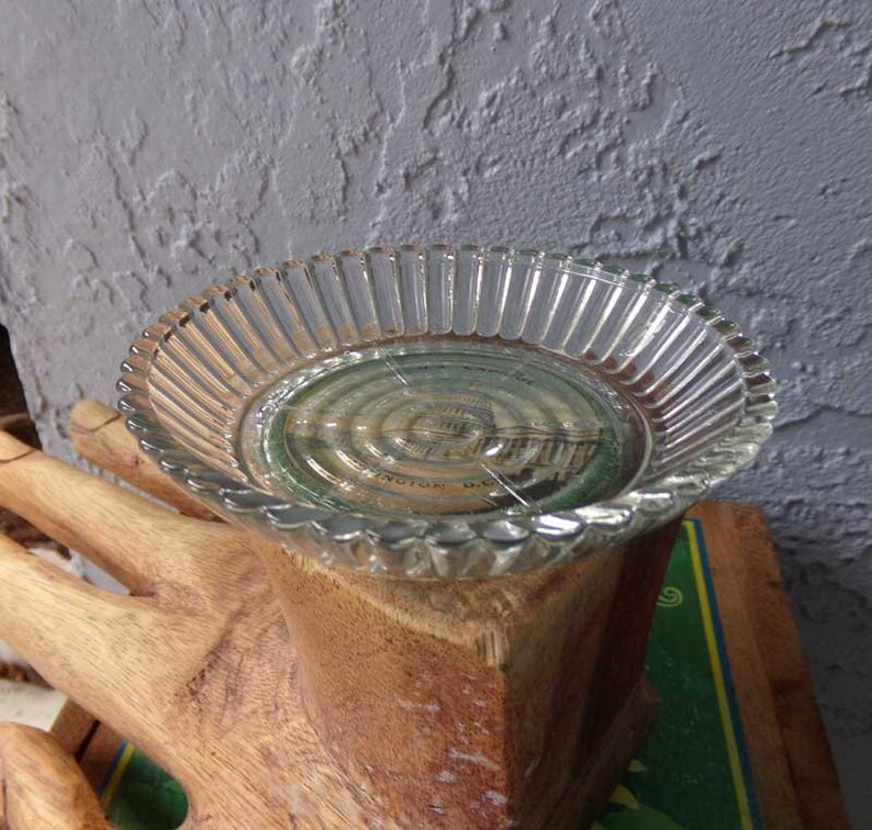May include: A clear glass dish with a fluted edge sits on top of a wooden hand sculpture. The dish has a circular design with a faint image of a building in the center.