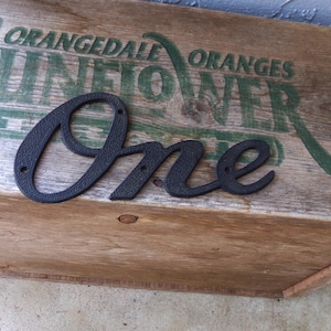 May include: A black metal sign in the cursive script font spelling the word One. The sign features a textured surface and three small circular holes for mounting purposes. The letters are connected in a single piece of dark, matte-finished metal.