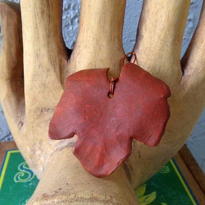 May include: A reddish-brown leaf-shaped pendant with a copper wire loop for hanging. The pendant is made of a textured material and is displayed on a wooden hand sculpture. The leaf has a naturalistic shape with detailed edges.