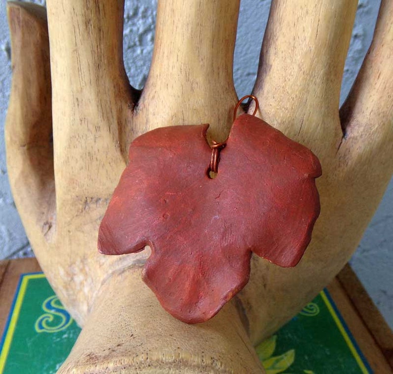 May include: A reddish-brown leaf-shaped pendant with a copper wire loop for hanging. The pendant is made of a textured material and is displayed on a wooden hand sculpture. The leaf has a naturalistic shape with detailed edges.