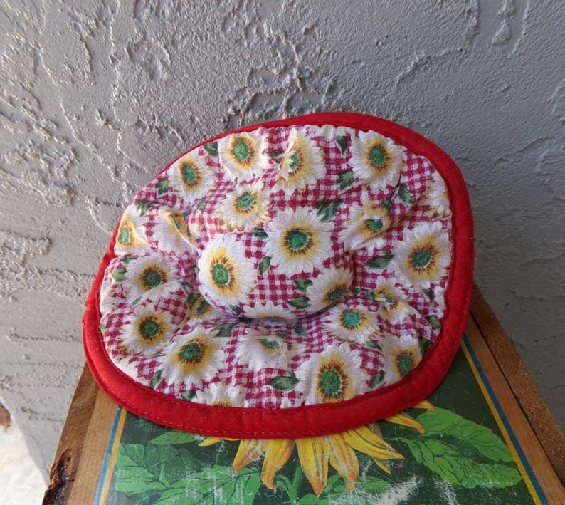 May include: Red and white pot holder with a sunflower and gingham pattern. The pot holder has a red border.