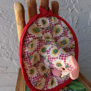 May include: A red and white fabric pot holder with a floral pattern and a red trim. The pot holder is shaped like a hand with five fingers and is sitting on a wooden surface.