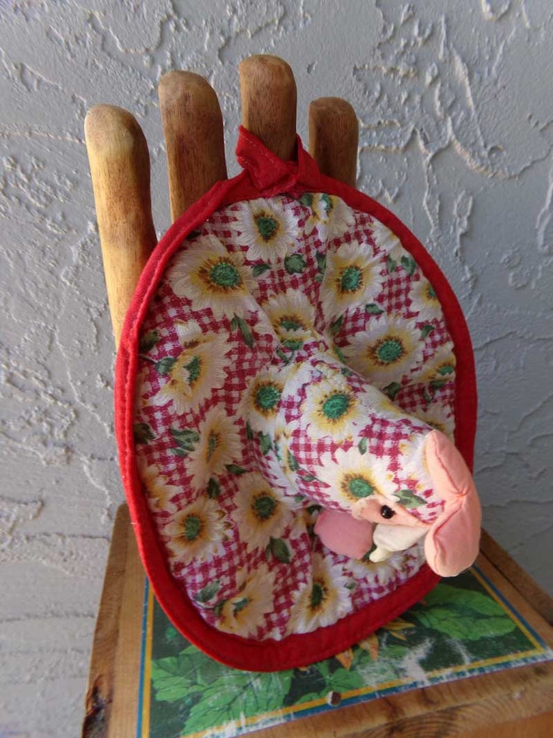 May include: A red and white fabric pot holder with a floral pattern and a red trim. The pot holder is shaped like a hand with five fingers and is sitting on a wooden surface.
