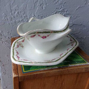 May include: White porcelain gravy boat with a gold rim and pink floral design. The boat is on a matching saucer with a square shape.