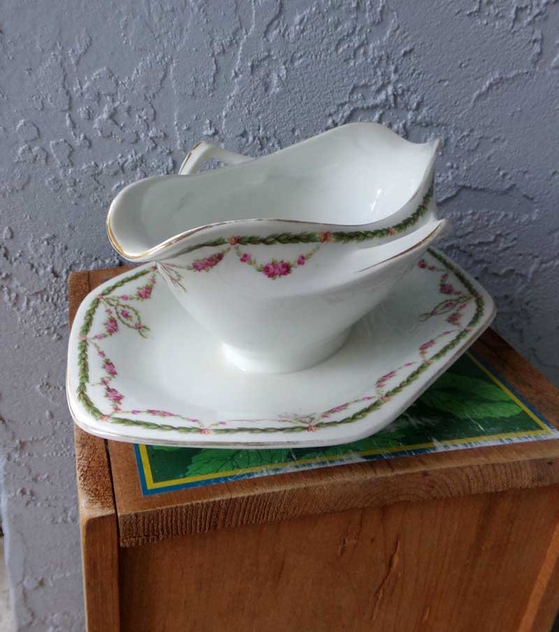 May include: White porcelain gravy boat with a gold rim and pink floral design. The boat is on a matching saucer with a square shape.