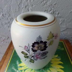 May include: White ceramic vase with a gold rim and floral design. The flowers are purple, pink, and green.