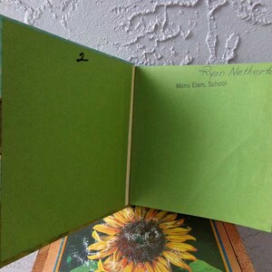 May include: A green book with a sunflower on the cover. The book is open to a blank page with the text "Ryan Netherton" and "Mims Elem. School" written on it. The number "2" is written on the other page.