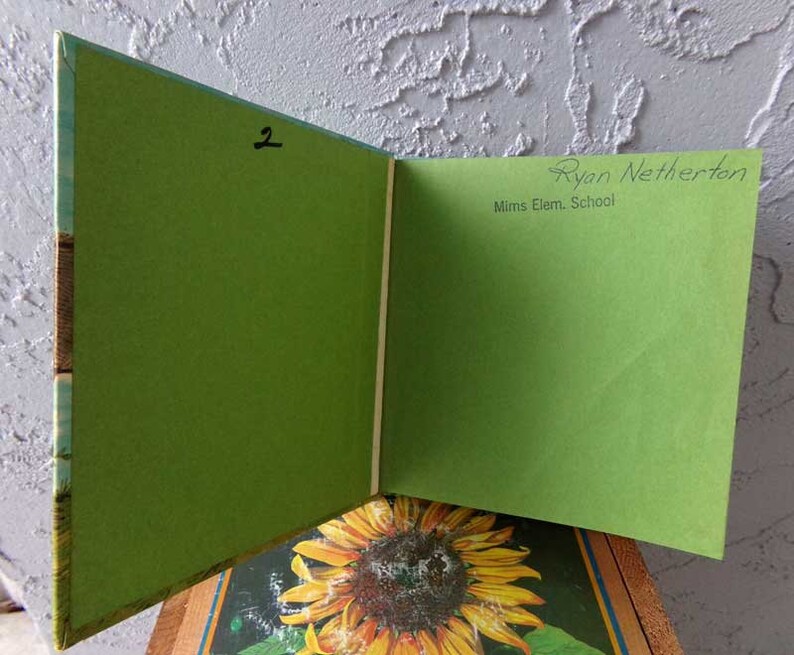 May include: A green book with a sunflower on the cover. The book is open to a blank page with the text "Ryan Netherton" and "Mims Elem. School" written on it. The number "2" is written on the other page.