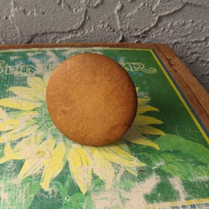 May include: A round, light brown wooden knob. The knob is set on a green and yellow painted surface with a sunflower design. The background is a textured gray wall.