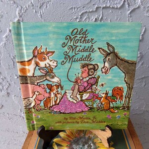 May include: A vintage children's book titled "Old Mother Middle Muddle" by Bill Martin Jr. with pictures by Don Madden. The cover features a colorful illustration of a woman knitting with various animals surrounding her.