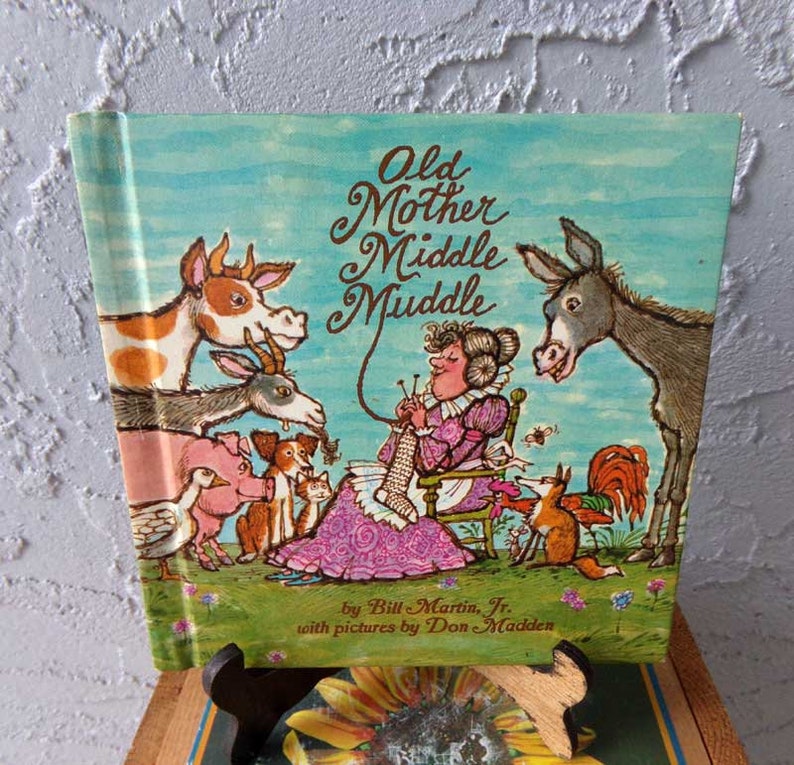 May include: A vintage children's book titled "Old Mother Middle Muddle" by Bill Martin Jr. with pictures by Don Madden. The cover features a colorful illustration of a woman knitting with various animals surrounding her.