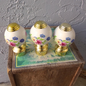 May include: Three decorative curtain finials. Each finial has a white, spherical body adorned with pink, blue, and yellow floral patterns. The finials have gold-colored bases and tops, adding a touch of elegance to the design.
