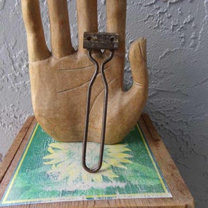 May include: A vintage metal bottle opener with a rusty finish. The opener has a simple design with a curved handle and a flat, rectangular head with two holes.