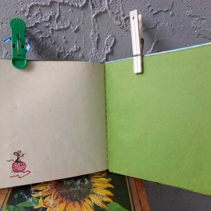May include: An open blank journal with a green cover and a small mouse illustration on the inside page. The journal is held closed with a green plastic clip and a white wooden clothespin.
