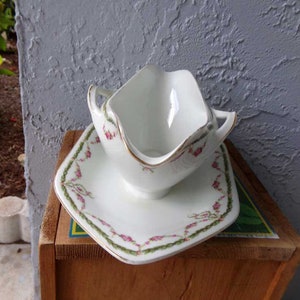 May include: White porcelain gravy boat with a gold trim and a floral design. The boat is on a matching saucer with a green and pink floral design.