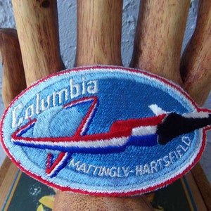 May include: A blue and white oval embroidered patch with the text "Columbia" and "Mattingly-Hartford" and a red, white, and blue space shuttle design.