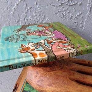 May include: A vintage children's book titled "Old Mother Middle Muddle" with colorful illustrations of animals and a woman in a pink dress. The book is on a wooden surface.