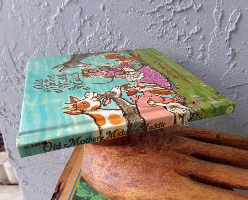 May include: A vintage children's book titled "Old Mother Middle Muddle" with colorful illustrations of animals and a woman in a pink dress. The book is on a wooden surface.