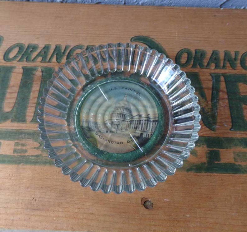 May include: A clear glass ashtray with a scalloped edge. The ashtray has a round, paper insert with a picture of the U.S. Capitol building in Washington D.C. The text "U.S. Capitol, Washington D.C." is printed on the insert.