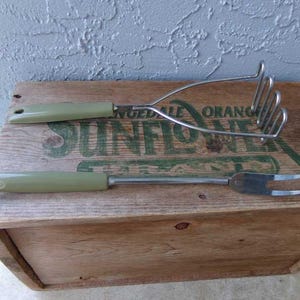 May include: A set of two vintage kitchen tools with green handles. The first tool is a potato masher with a silver metal head. The second tool is a fork with a silver metal head. Both tools are sitting on a wooden crate with the words "Sun Flower" printed on it.