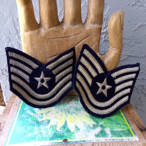 May include: Two vintage US Air Force technical sergeant rank insignia patches. The patches are blue with a white star and three silver stripes. The patches are on a wooden surface.