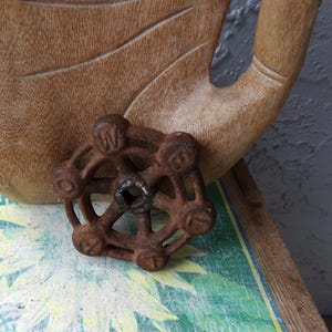 May include: A rusty, cast iron valve handle with a circular design and a central hole. The handle is resting on a wooden surface with a carved hand and a painted floral pattern.