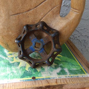 May include: A rusty, blue and brown metal valve handle with a star-like design. The handle has a square center and is set against a wooden hand sculpture and a colorful background.