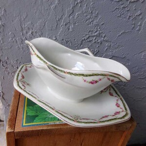 May include: White porcelain gravy boat with a matching saucer. The boat has a delicate floral design with pink roses and green vines. The saucer has a matching design around the edge.