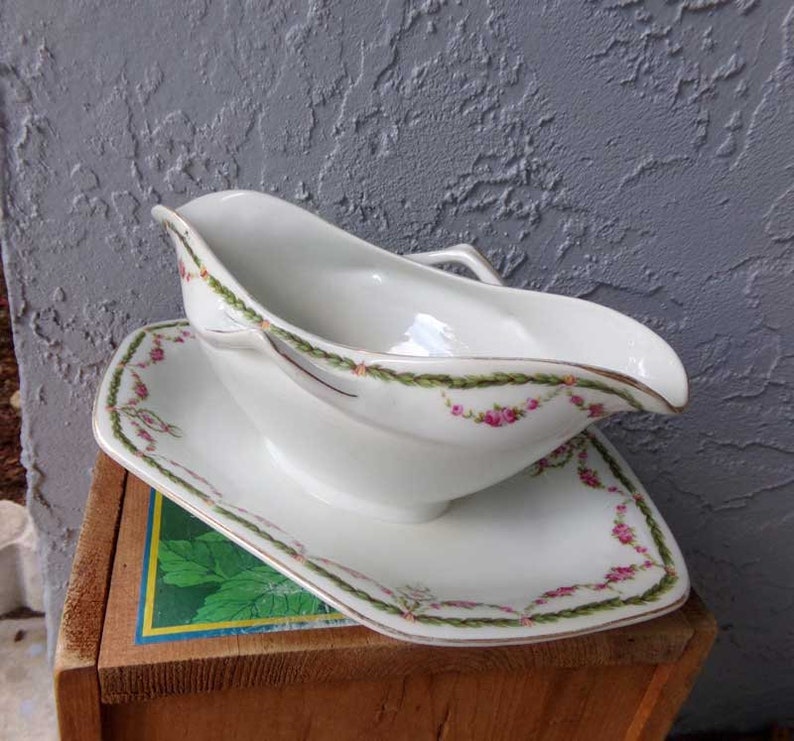May include: White porcelain gravy boat with a matching saucer. The boat has a delicate floral design with pink roses and green vines. The saucer has a matching design around the edge.