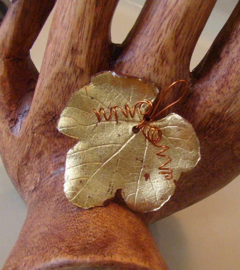 May include: A gold-colored leaf-shaped brooch with copper wire detailing. The brooch has a textured surface and is attached to a wooden hand-shaped display. The wire is coiled and adds a decorative element to the leaf.