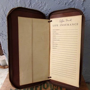 May include: A brown leather zippered binder with a cream-colored page inside. The page has the title "Coffee Fund LIFE INSURANCE" and a list of fields to fill in, including "Name of Insured", "Company", "Policy Number", "Amount", "Premium", "Premium Payable", "Plan", "Date of Issue", "Age at Issue", and "Beneficiary".